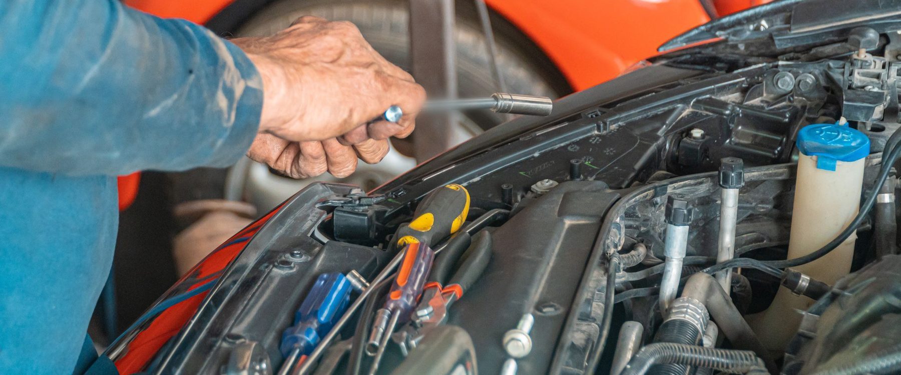Mão de mecânico aperta uma peça no cofre do motor com chave de soquete, ao lado de chaves de fenda, com um carro laranja ao fundo